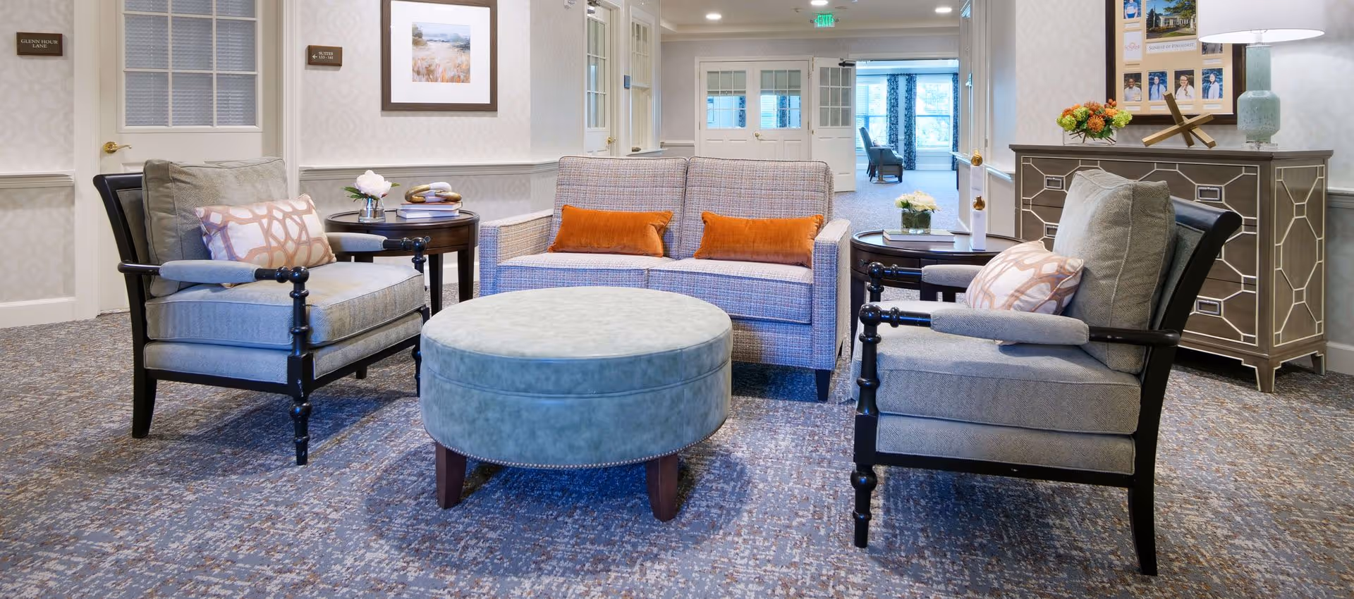 A bright lounge area with two upholstered armchairs, a loveseat, round ottoman, and side tables in a senior living facility.
