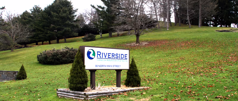 Outdoor view of a grassy hillside with a sign that reads 'Riverside Center for Rehabilitation & Nursing, 90 North Main Street' surrounded by small evergreen shrubs and trees in the background.