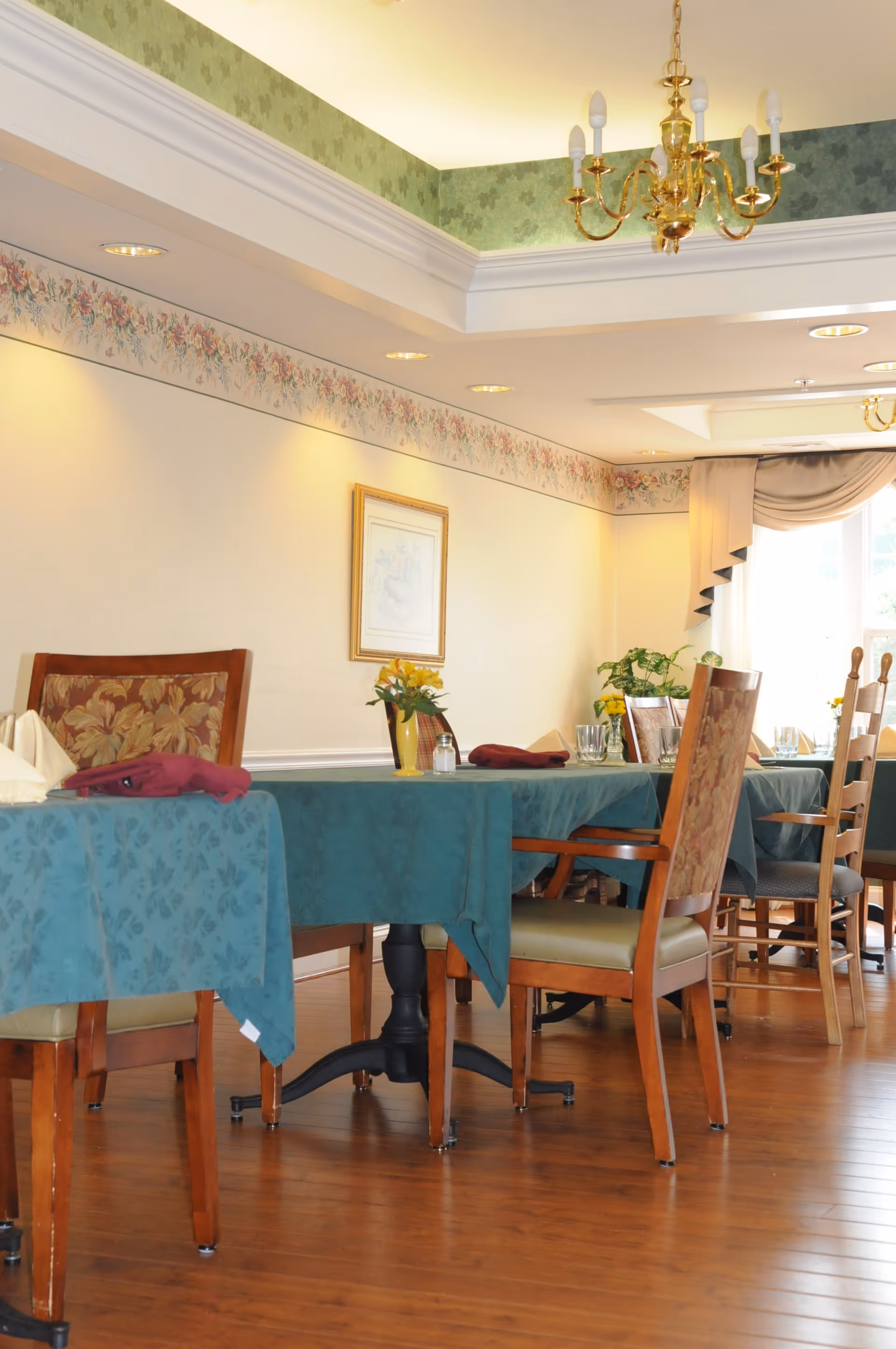 A dining room with wooden floors and tables covered with green tablecloths. Chairs with floral upholstery are arranged around the tables. The room has floral wallpaper borders near the ceiling, a chandelier, framed artwork on the wall, and windows with curtains allowing natural light to enter.