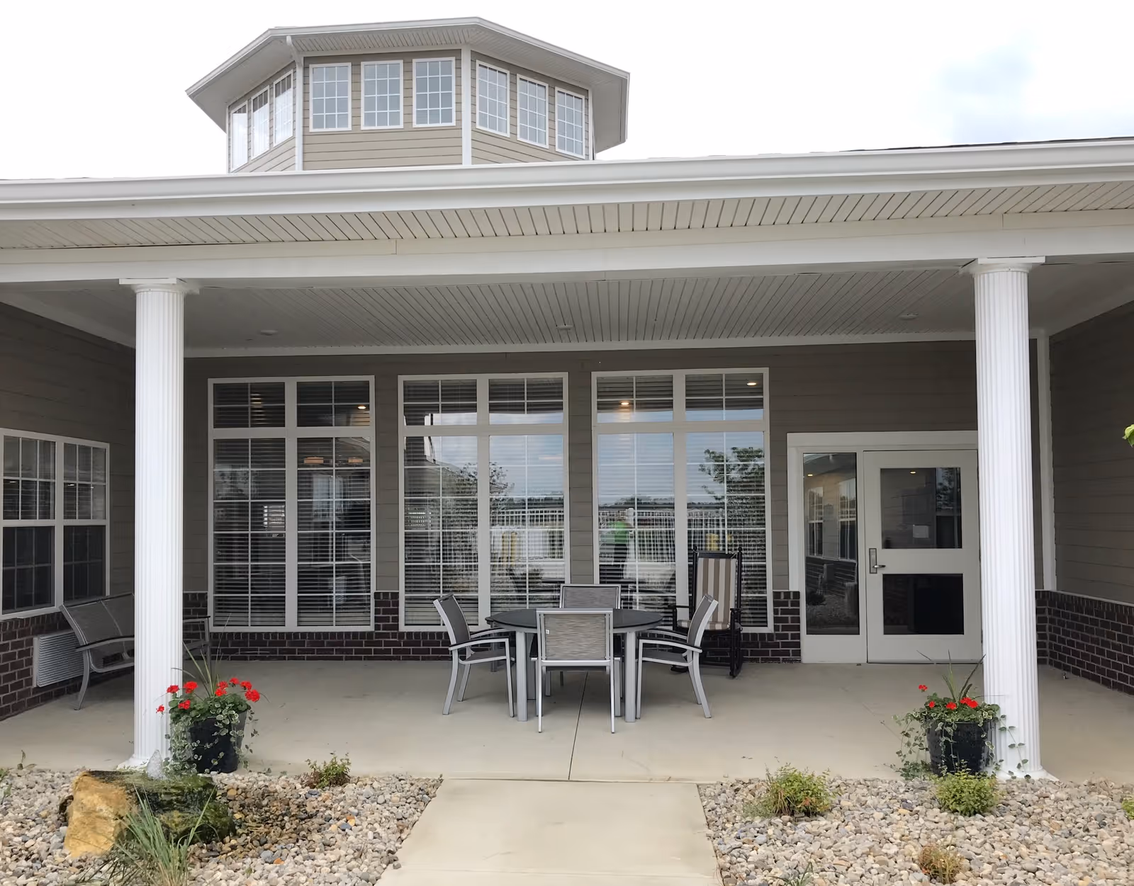 Covered outdoor patio area at The Springs of Lima with white columns, a round table with four chairs, two benches, potted plants with red flowers, and large windows with white blinds behind the seating area.