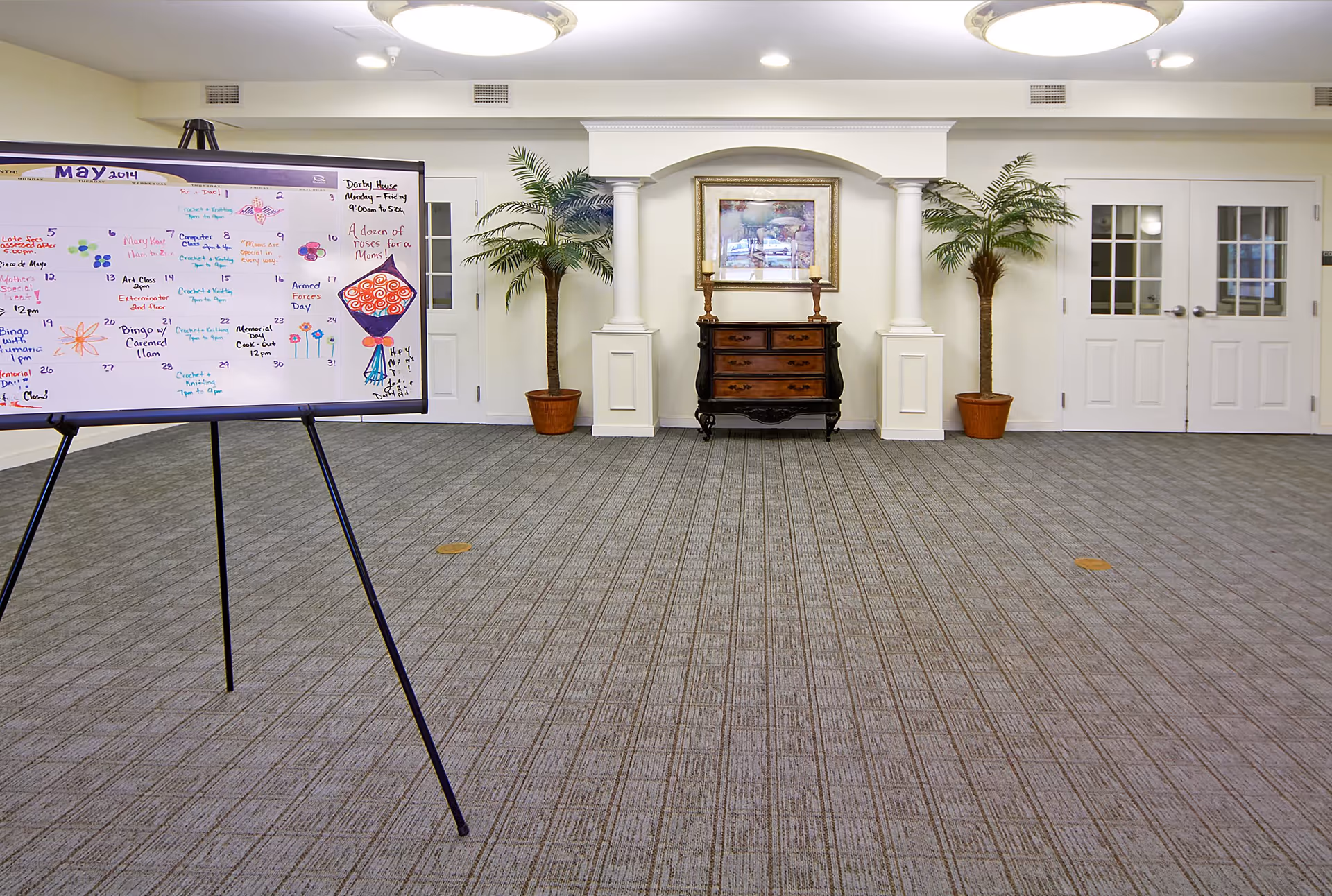 Interior room with a patterned carpet floor, two potted palm plants on either side of a decorative wooden chest with a framed picture above it, and a whiteboard on a tripod stand displaying a May 2014 calendar with handwritten notes and drawings.