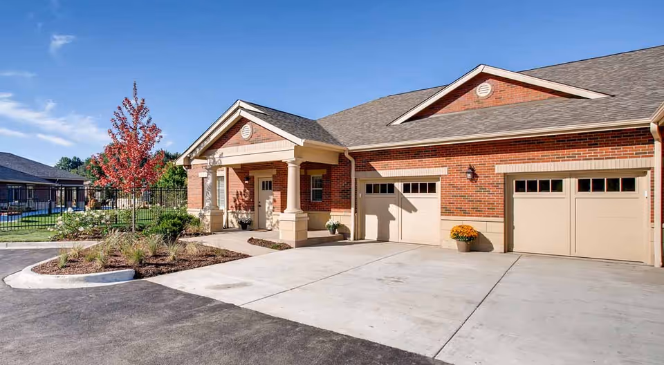 Exterior view of a brick building with a covered entrance supported by columns, two garage doors, a small landscaped area with plants and a tree with red leaves, and a clear blue sky.