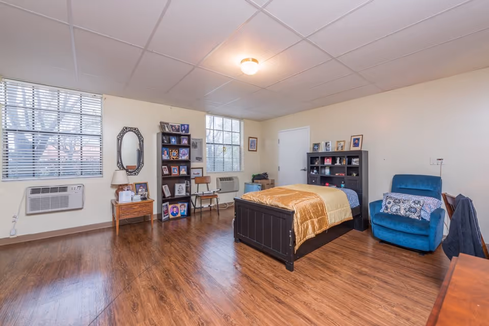 A spacious bedroom with wooden flooring, a single bed with a golden blanket, a blue armchair with a patterned pillow, a wooden side table with a lamp and framed photos, two windows with blinds, a bookshelf filled with framed photos, and a mirror on the wall.