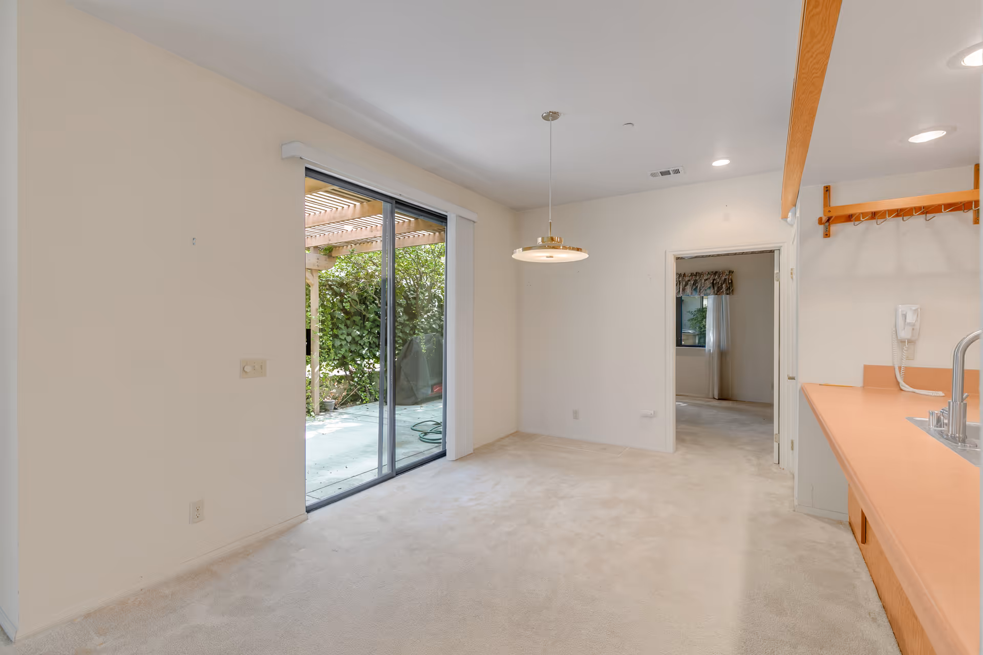 Empty interior room with beige carpet, a sliding glass door leading to an outdoor patio with greenery, a hanging ceiling light, and a countertop with a sink and wall-mounted phone on the right side. There is an open doorway leading to another room with a window and curtains.