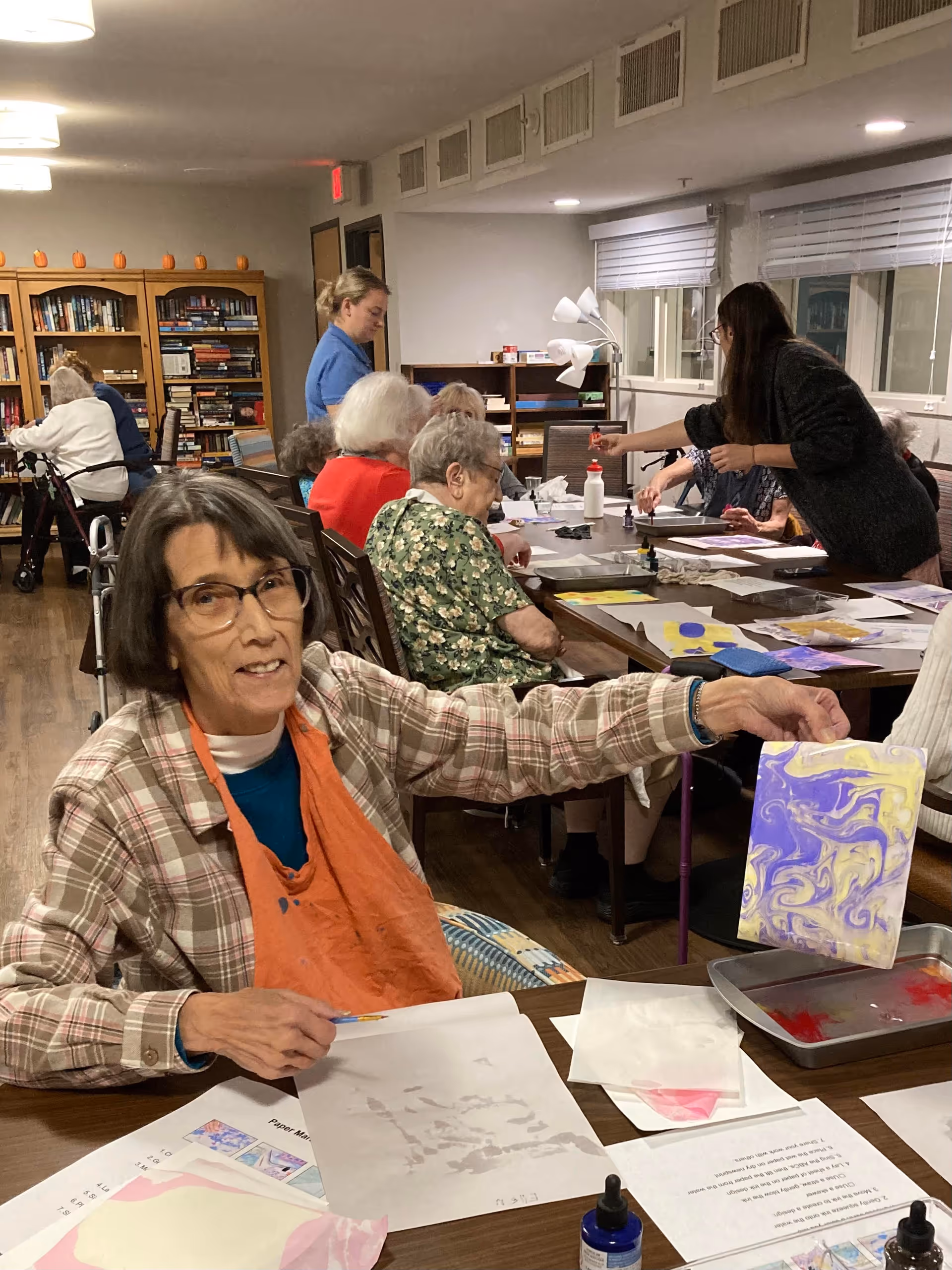 An elderly woman wearing glasses and an orange apron is sitting at a table in a senior living facility art room, holding up a piece of paper with a purple and yellow marbled design. Other elderly residents and staff are engaged in art activities in the background, with bookshelves and windows visible in the room.