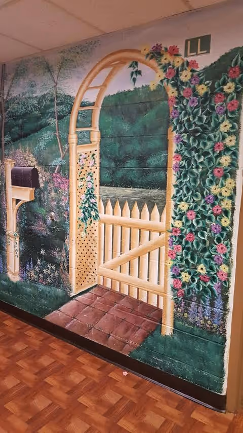 A mural painted on an interior wall depicting a garden scene with a wooden arched gate, a white picket fence, colorful flowers climbing the arch, a mailbox on a post, and green trees and bushes in the background. The floor in front of the mural has a wood-patterned surface.