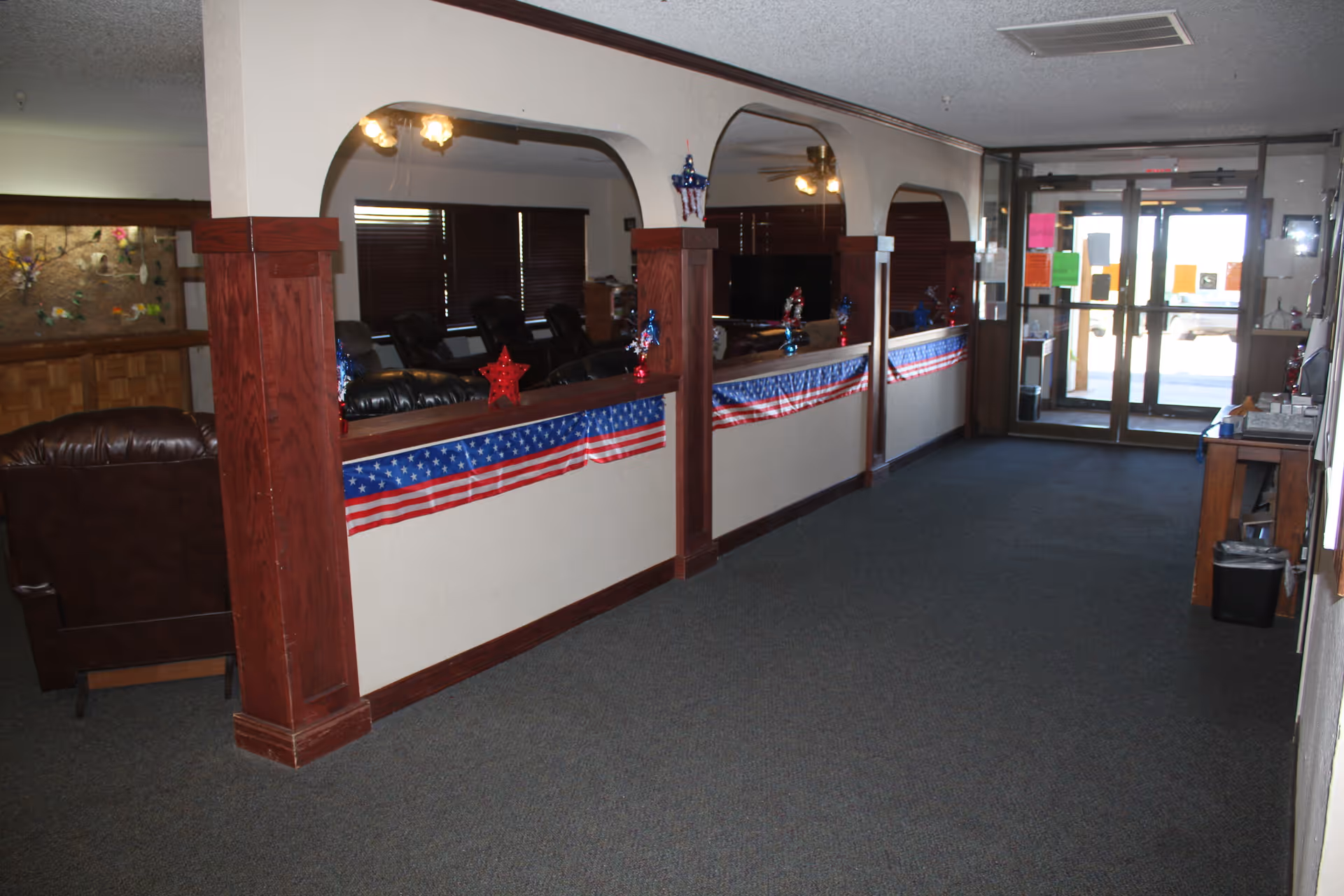 Interior hallway of a senior living facility decorated with patriotic red, white, and blue banners and star ornaments. The hallway features wooden pillars and arches separating it from a lounge area with leather recliners and a television. Double glass doors are visible at the end of the hallway.