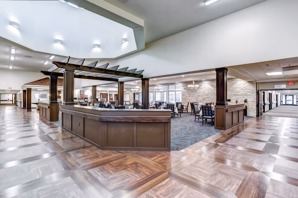 Spacious senior living dining and common area with a central wood service counter, pergola-style columns, tables and chairs, and polished wood floors.