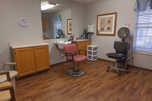Interior of a hair salon room with wooden flooring, a red salon chair in the center, a black salon chair with a hair dryer hood on the right, wooden cabinets, a wall-mounted sink with a mirror above it, a clock on the wall, and a window with blinds and a blue valance.