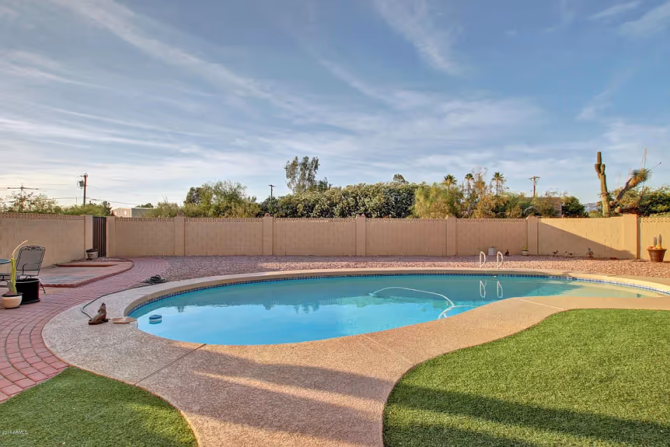 Outdoor view of a kidney-shaped swimming pool surrounded by a concrete deck and artificial grass, enclosed by a beige brick wall with some potted plants and trees visible beyond the wall under a partly cloudy sky.
