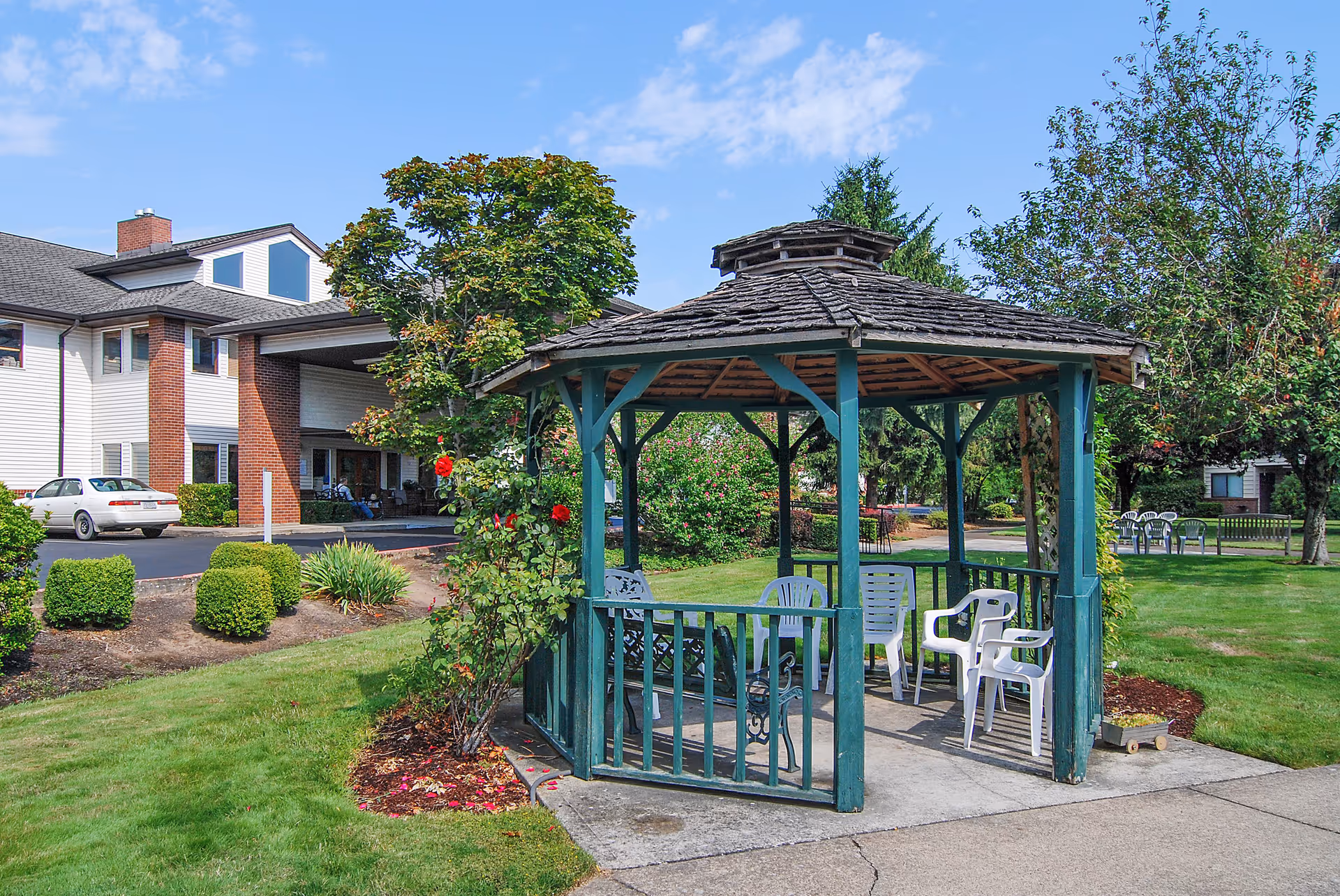 A green wooden gazebo with white plastic chairs on a grassy lawn near the entrance of a multi-story senior living building.