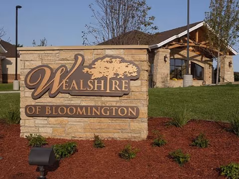 Stone sign with the text 'Wealshire of Bloomington' in front of a building with a sloped roof and large windows, surrounded by grass, small plants, and mulch.