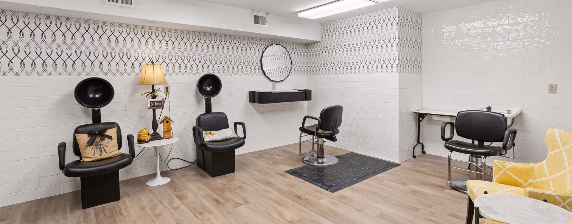Interior of a salon area in a senior living facility with two black vintage hair dryer chairs, a small white side table with a lamp and decorative items, a black salon chair in front of a wall-mounted shelf and mirror, and another black chair at a small table. The room has light wood flooring, white walls with a patterned wallpaper border near the ceiling, and a yellow patterned armchair partially visible on the right.