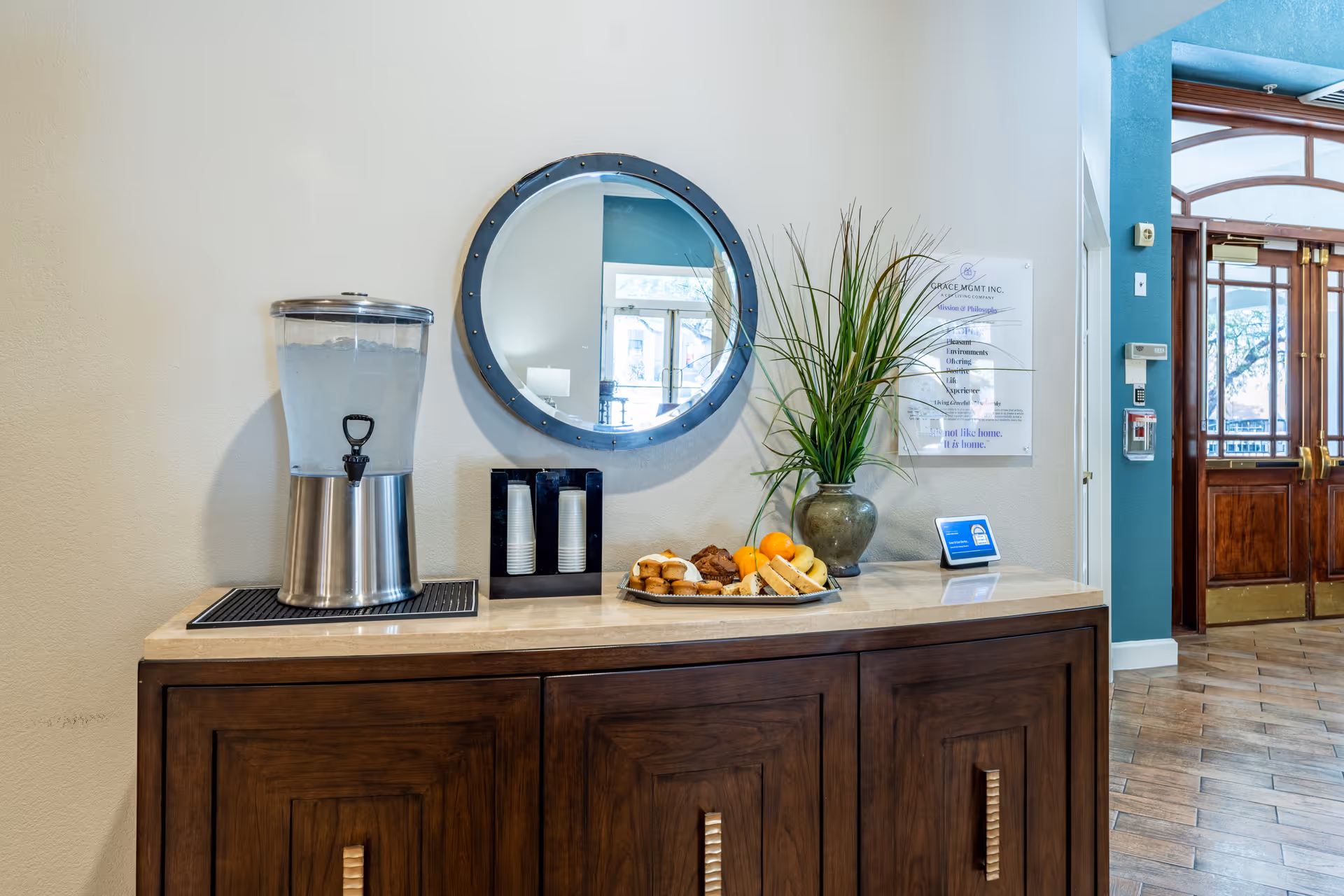 A wooden sideboard with a water dispenser, a holder with stacked disposable cups, a plate of assorted pastries and fruits, and a vase with tall green plants. Above the sideboard is a round mirror reflecting part of the room. To the right, there is a wall-mounted sign and a small digital device on the countertop. The background shows a doorway with wooden double doors and a tiled floor.