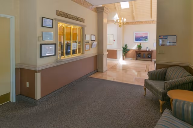 Interior view of a senior living facility lobby area with a reception window on the left wall, framed certificates and notices around it, a carpeted floor leading to a tiled area with a small table and chairs, a chandelier hanging from a high ceiling, and a framed picture on the far wall.