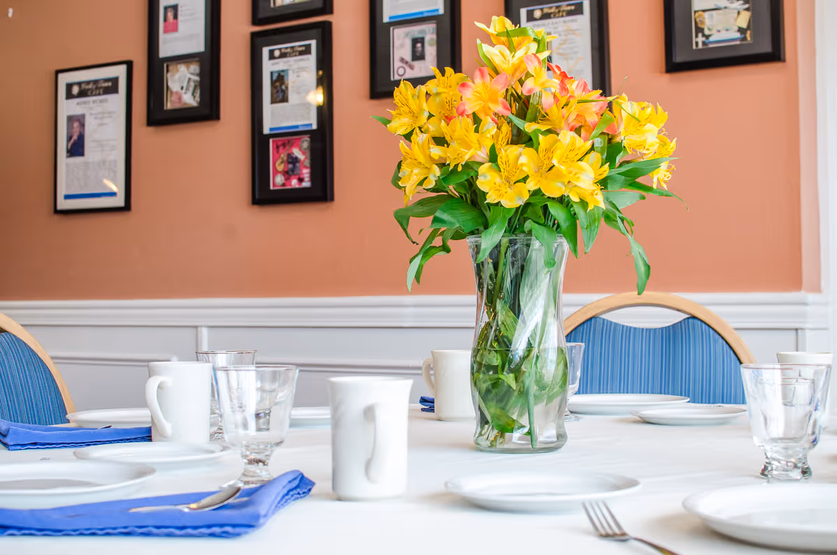 A dining table set with plates, cups, glasses and a vase of yellow flowers in a room with framed photos on the wall.