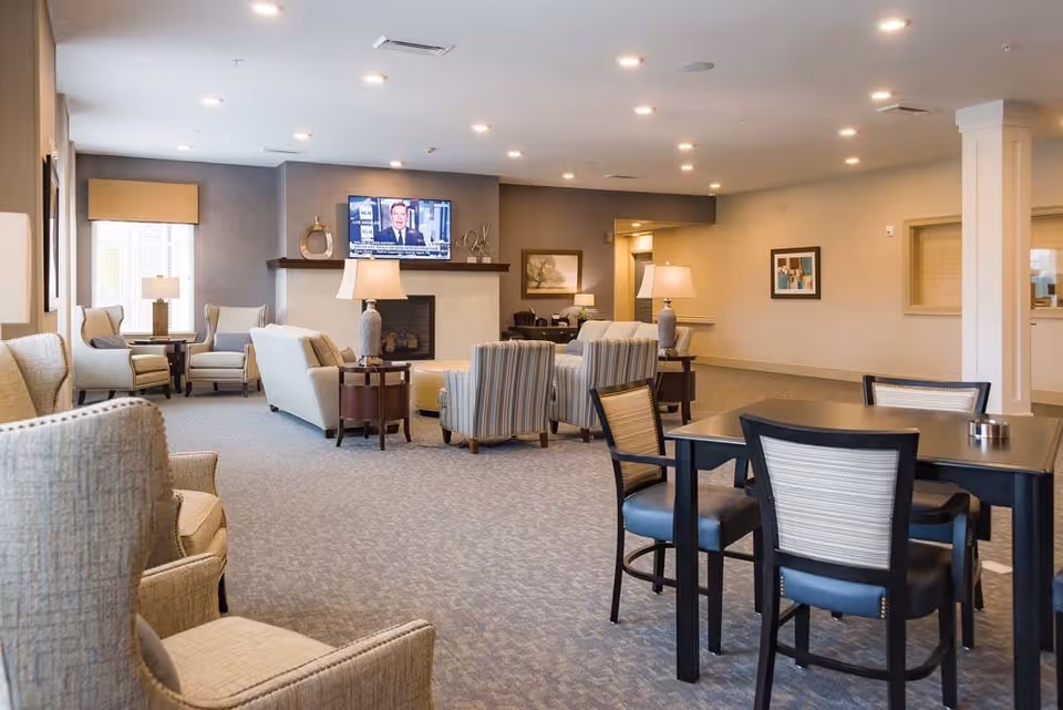 A spacious and well-lit living room area in a senior living facility with multiple armchairs and sofas arranged around a fireplace and a wall-mounted television. There are also tables with chairs in the foreground and lamps providing warm lighting. The walls are decorated with framed artwork.