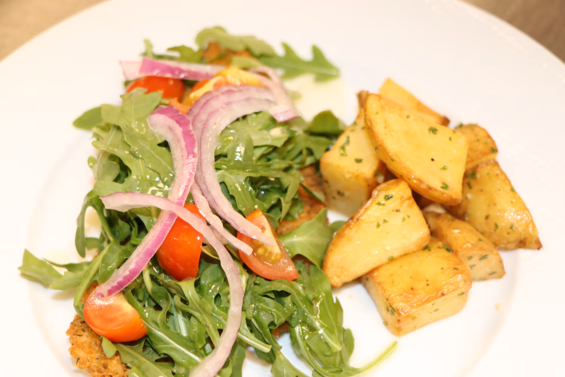A plate with an arugula salad topped with cherry tomatoes and red onion slices served alongside roasted potato wedges.