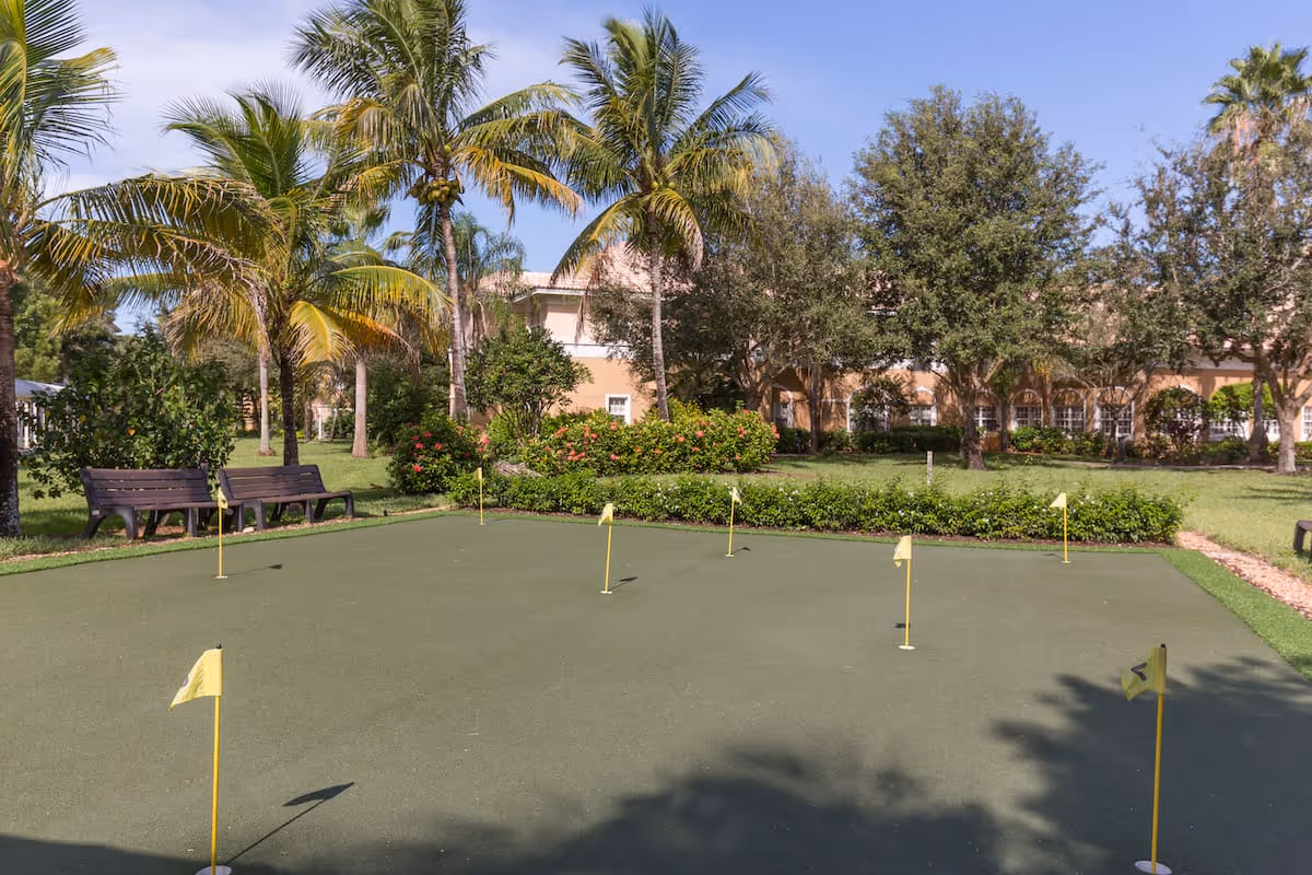 Outdoor putting green with several small yellow flags on poles, surrounded by palm trees, bushes, and benches, with a building visible in the background under a clear sky.