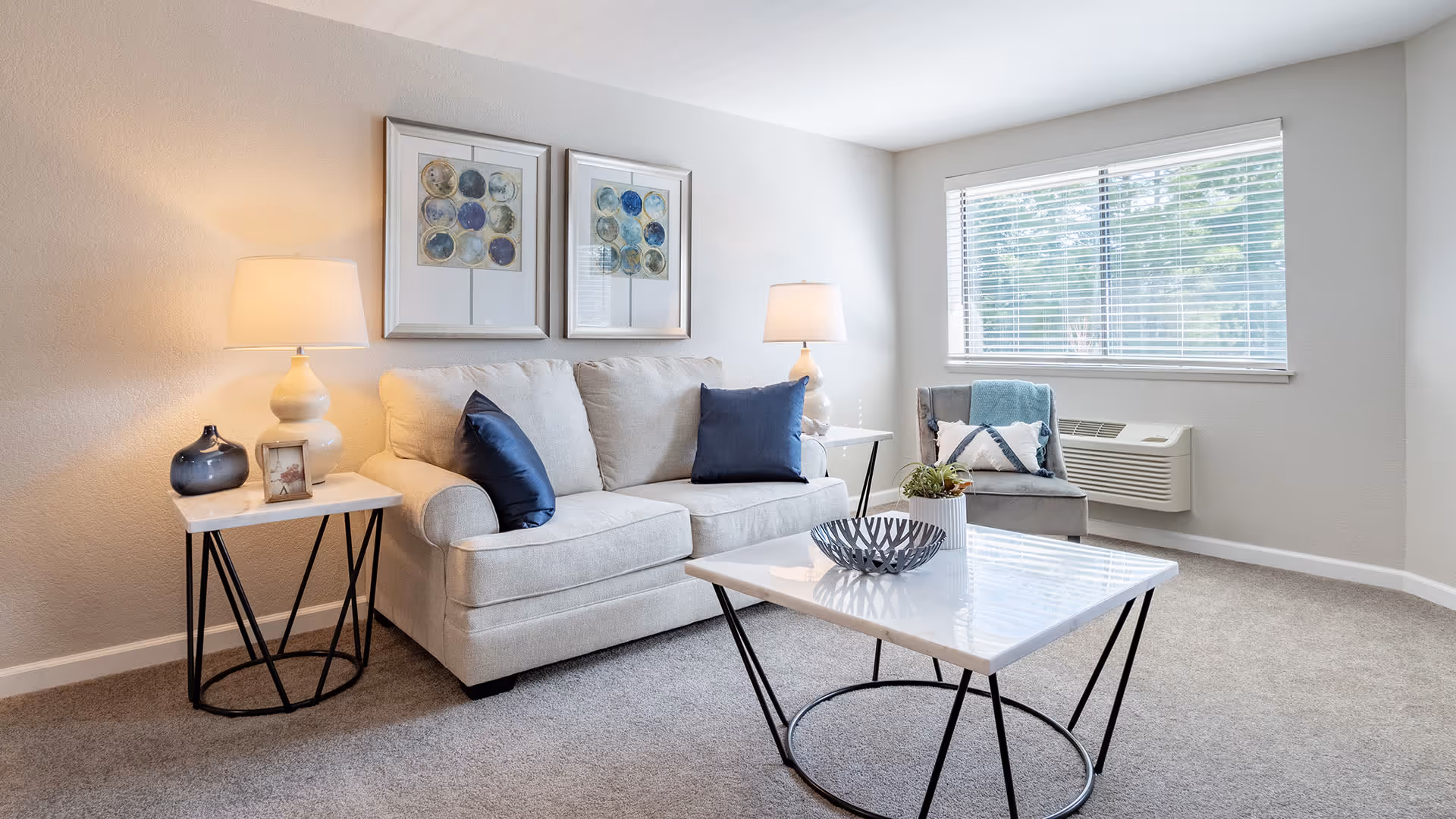 A bright living room with a beige sofa adorned with two dark blue pillows, a gray armchair with a blue throw and pillow, two side tables each with a lamp, a decorative bowl on a white coffee table, and two framed abstract artworks on the wall. A window with blinds lets in natural light, and a wall-mounted air conditioning unit is visible below the window.