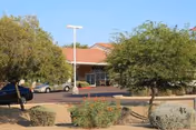 Exterior view of a single-story building with a red-tiled roof, surrounded by trees and shrubs, with a parking area in front containing several cars under a clear sky.