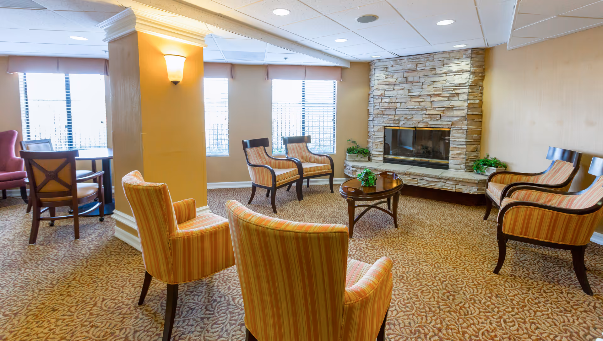 A cozy senior living facility common area with several striped upholstered armchairs arranged around a wooden coffee table. The room features a stone fireplace, beige walls, patterned carpet, and large windows with blinds allowing natural light to enter.