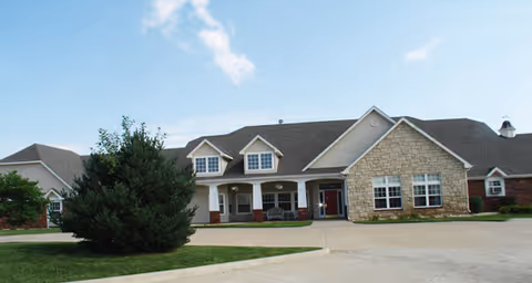 Exterior view of a single-story senior living facility building with a combination of stone and siding facade, multiple windows, a covered front porch with seating, and a paved driveway in front. The sky is clear with a few clouds.