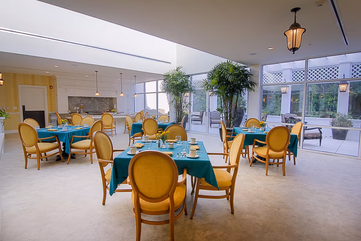 Bright dining room with round tables set with teal tablecloths and yellow upholstered chairs, large windows and potted plants.