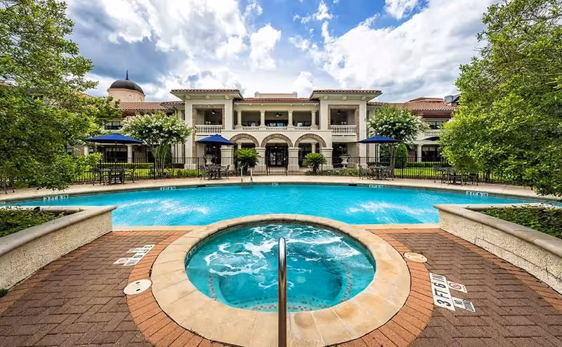 Outdoor view of a senior living facility featuring a swimming pool and a circular hot tub in the foreground, surrounded by brick paving and greenery. The building in the background has a Mediterranean architectural style with arches, balconies, and a tiled roof under a partly cloudy sky.