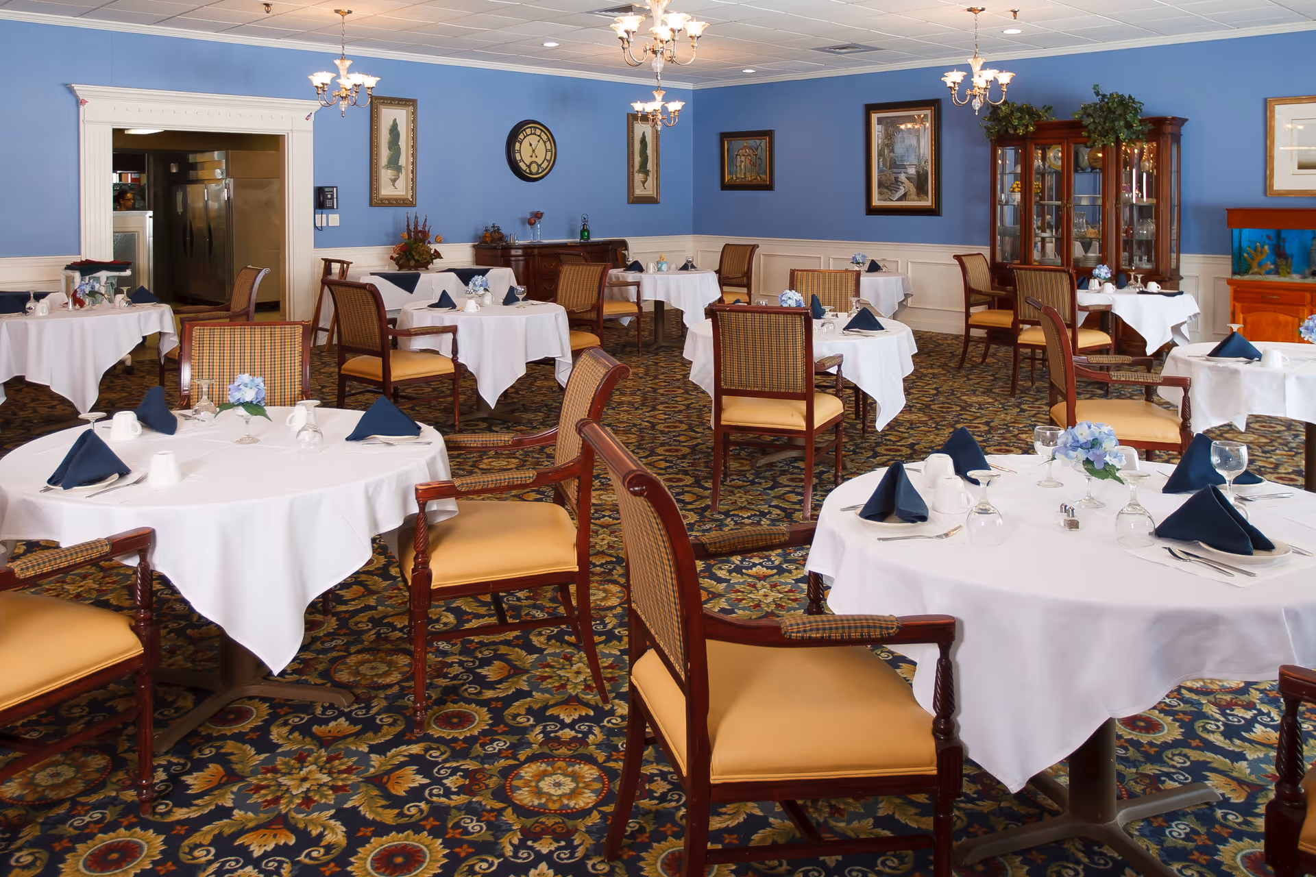 A dining room with multiple round tables covered with white tablecloths, each set with navy blue folded napkins, glassware, and floral centerpieces. The room has blue walls adorned with framed artwork and a clock, a patterned carpet, wooden chairs with cushioned seats, and chandeliers hanging from the ceiling. A china cabinet and a fish tank are visible in the background.