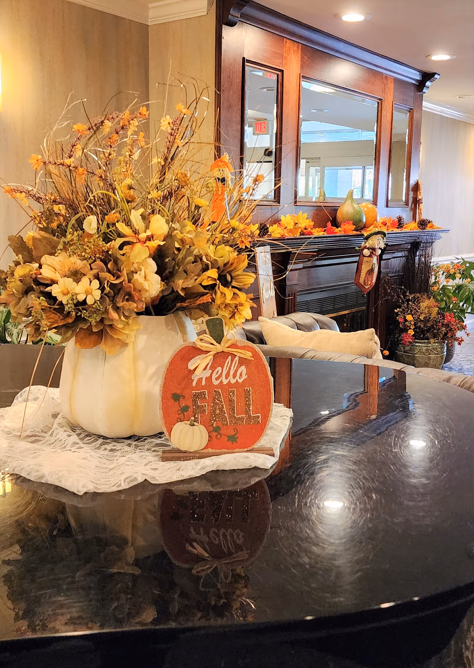 A cozy indoor setting featuring a dark polished table with a white pumpkin-shaped vase filled with autumn flowers and foliage. In front of the vase is a small decorative sign that reads 'Hello FALL'. Behind the table is a dark wooden fireplace mantel decorated with fall-themed items including pumpkins and garlands. A large mirror is mounted above the mantel, reflecting part of the room and an exit sign. The overall ambiance is warm and inviting, decorated for the fall season.