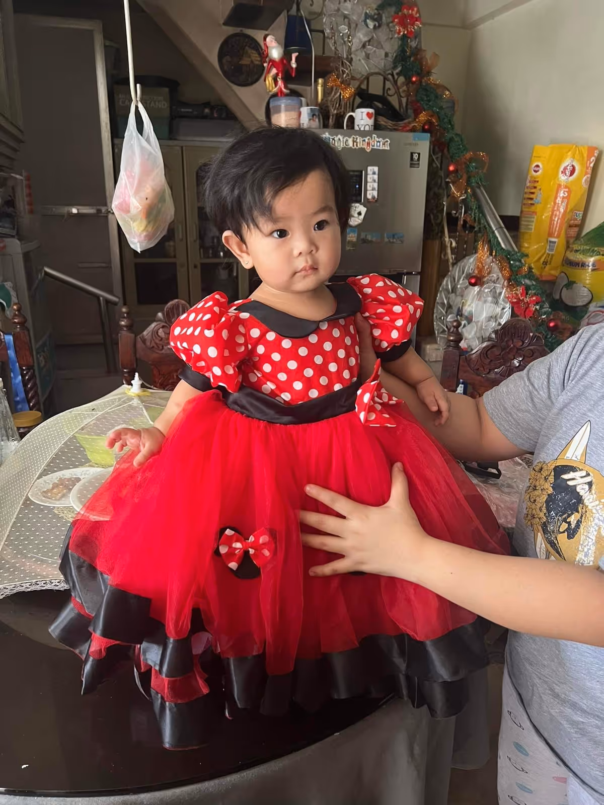 A young child wearing a red and black polka dot dress with puffed sleeves stands on a table while being supported by an adult. The background shows a kitchen area with a refrigerator, some hanging decorations, and various household items.