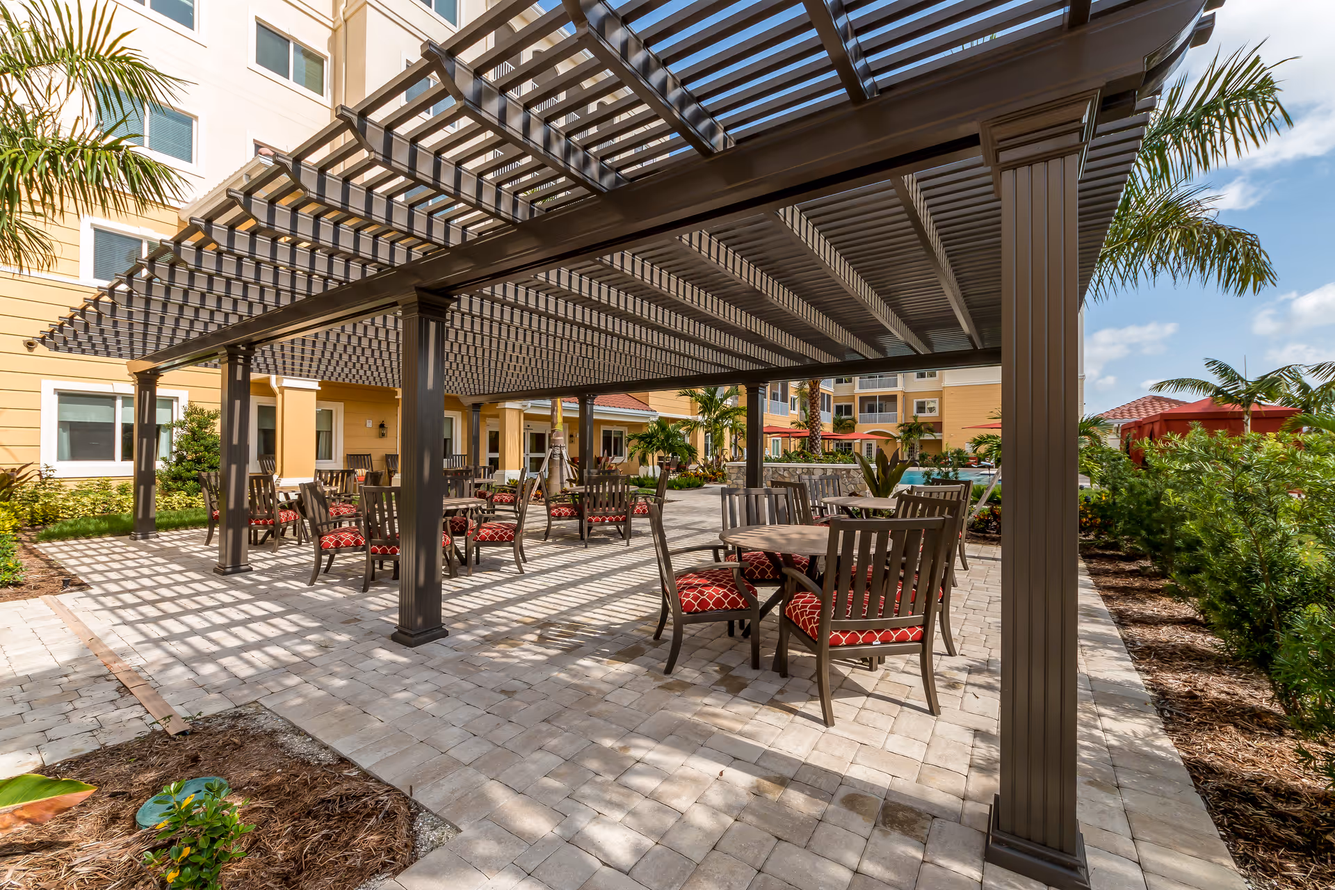 Outdoor patio area at Discovery Village At The Forum with a large pergola providing shade over several tables and chairs with red patterned cushions. The patio is paved with stone tiles and surrounded by landscaped greenery and palm trees, with the building visible in the background under a partly cloudy sky.