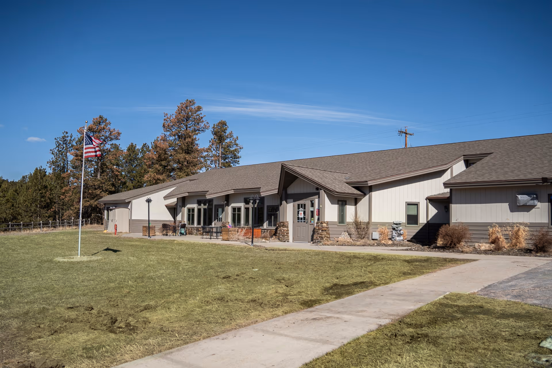Exterior view of a single-story assisted living facility building with a gray roof and beige siding, a concrete walkway leading to the entrance, an American flag on a flagpole in the grassy front yard, and trees in the background under a clear blue sky.