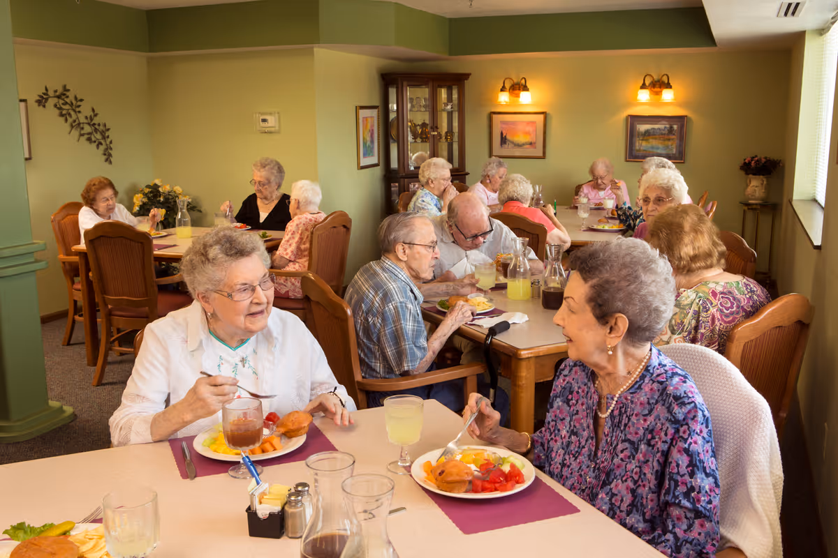 A group of elderly people sitting at tables in a dining room, enjoying meals and drinks while engaging in conversation. The room has green walls, framed paintings, and warm lighting fixtures. The tables are set with plates of food, glasses, and pitchers of beverages.