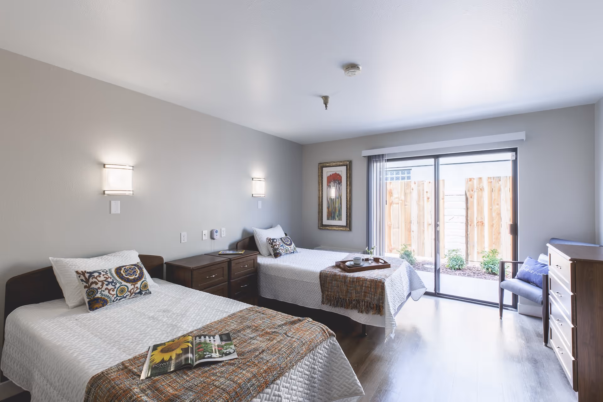 A bright bedroom with two single beds, each with white bedding and decorative pillows. There are two wall-mounted lights above the beds, a wooden nightstand between them, and a wooden dresser on the right side. A sliding glass door leads to an outdoor area with a wooden fence and small plants. A framed artwork hangs on the wall near the door, and a chair with a cushion is placed next to the dresser.