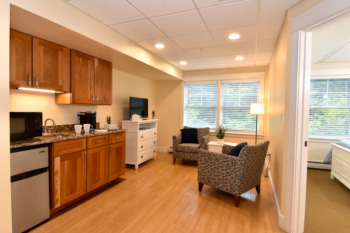 A cozy interior space featuring a small kitchenette with wooden cabinets, a microwave, mini fridge, and granite countertop. Adjacent to the kitchenette are two patterned armchairs with dark cushions, a small white side table with a plant, and a floor lamp near large windows with blinds. A white dresser with a TV on top is positioned against the wall. To the right, an open doorway reveals a bedroom with a white bed frame and large windows.