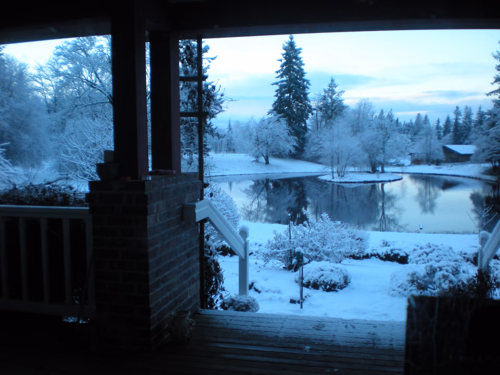 View from a covered porch overlooking a snowy landscape with a pond surrounded by snow-covered trees and bushes under a cloudy sky.