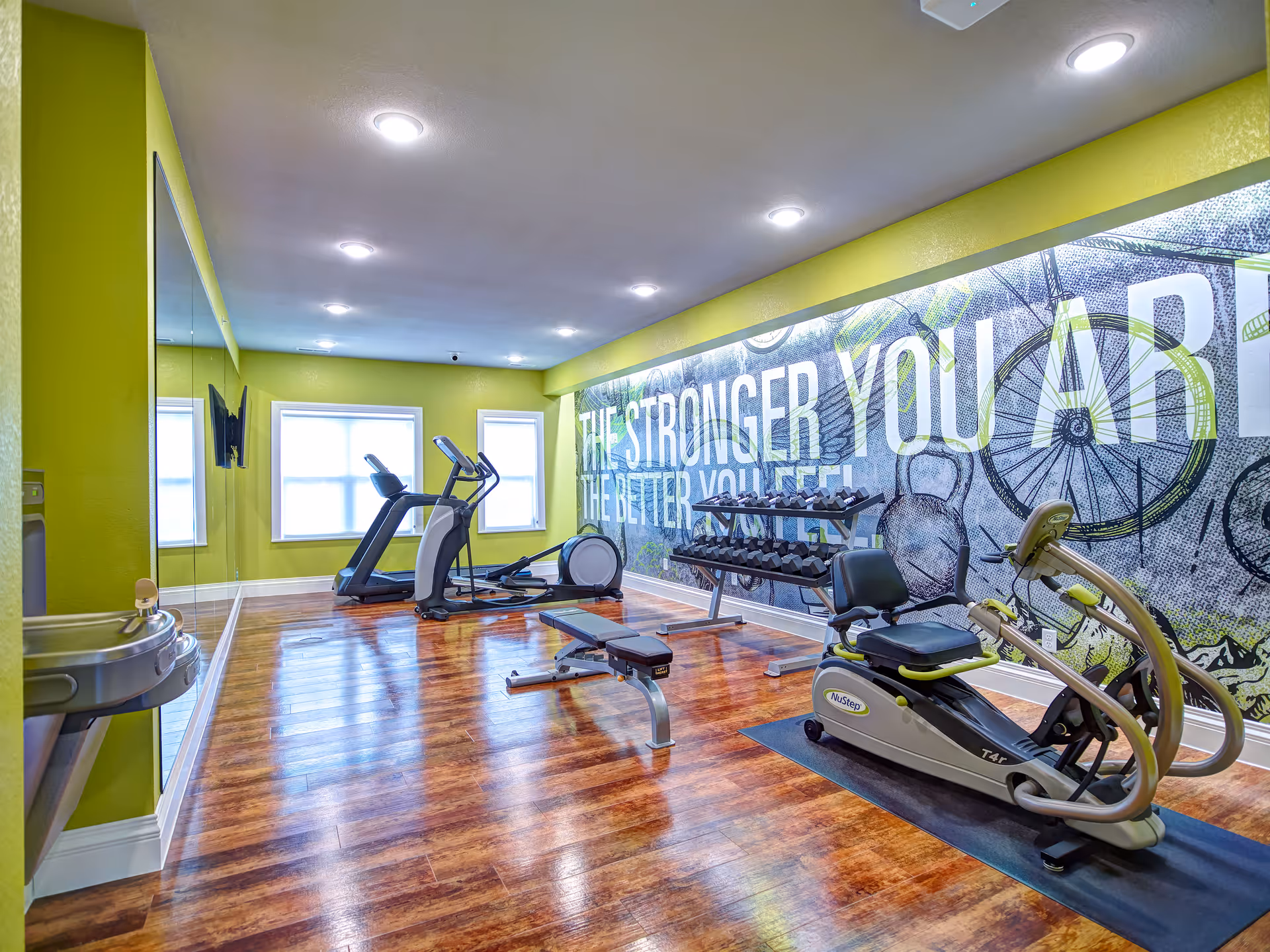 A fitness room with wooden flooring and bright green walls. The room contains exercise equipment including a treadmill, elliptical machine, recumbent bike, and a bench in front of a rack of dumbbells. A large wall mural with the text 'THE STRONGER YOU ARE THE BETTER YOU FEEL' is visible on one side. There are two windows letting in natural light and a water fountain mounted on the left wall.