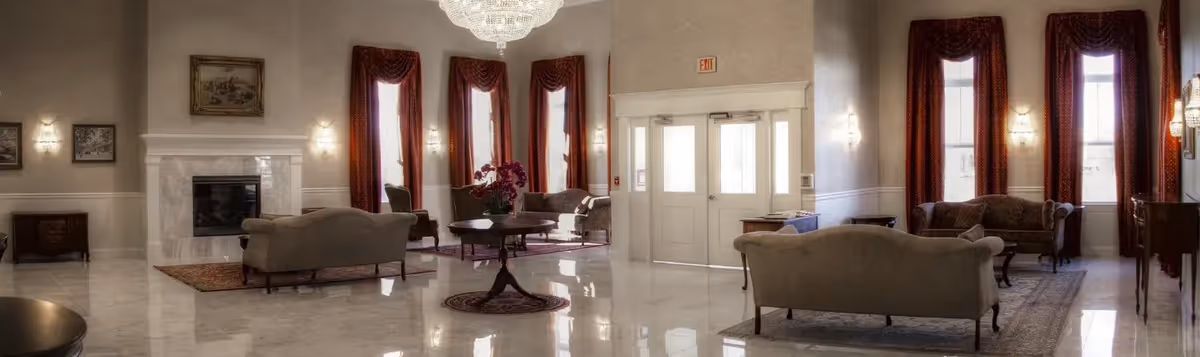 A spacious and elegant living room area with polished marble floors, multiple seating arrangements including sofas and armchairs, a white fireplace, large windows with red curtains, and a chandelier hanging from the ceiling. The room has a classic and refined decor style.