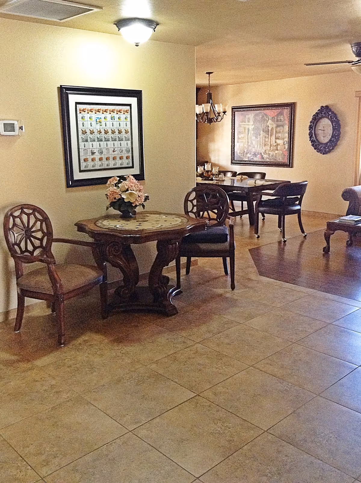 Interior view of a senior living facility showing a small wooden table with a floral centerpiece and two chairs in the foreground. In the background, there is a larger dining table with chairs, a chandelier, a framed painting on the wall, and a decorative wall clock. The floor is tiled and the walls are painted beige.