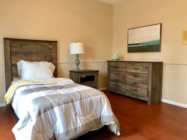 A simple bedroom with a single bed featuring a wooden headboard, white pillow, and a gray and white comforter with a yellow stripe. Next to the bed is a wooden nightstand with a gray lamp on it. Across from the bed is a wooden dresser with multiple drawers and a decorative bowl on top. A framed abstract painting hangs on the beige wall above the dresser. The room has wooden flooring and beige walls with white trim.