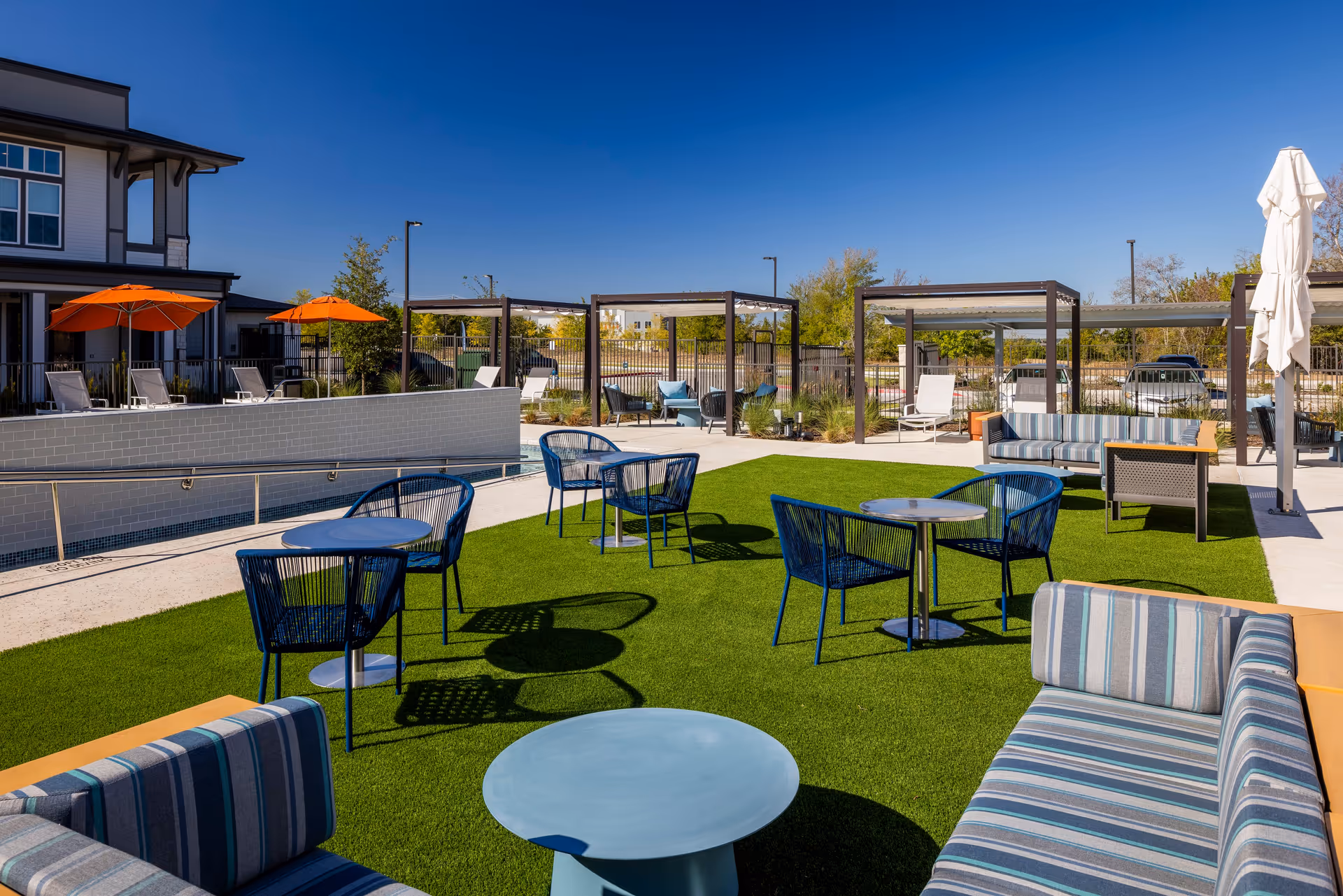 Outdoor seating area at Amberlin at The Station featuring blue chairs and tables on artificial grass, striped cushioned sofas, pergolas with additional seating, orange umbrellas, and a clear blue sky.