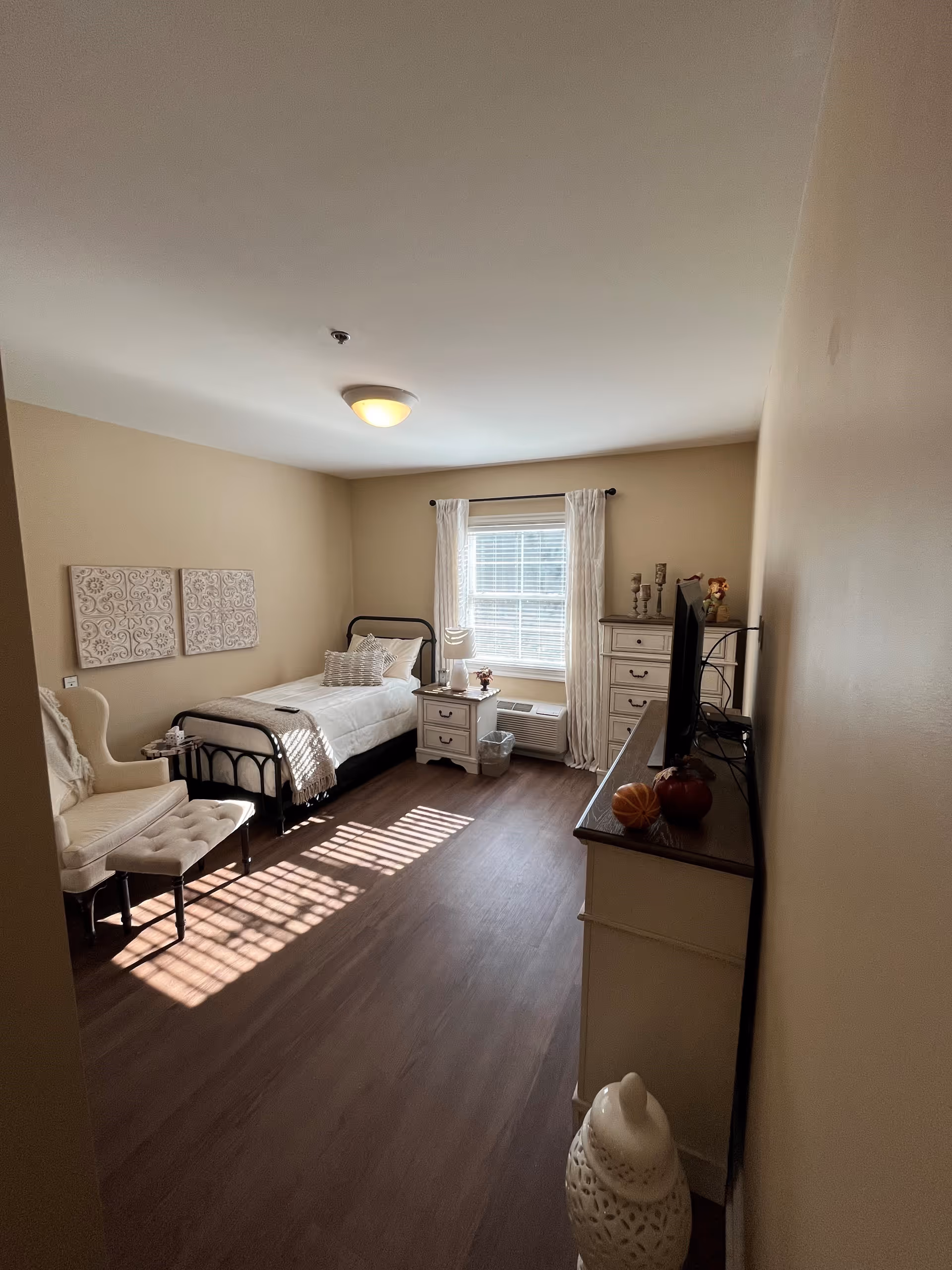 A bright, neatly arranged bedroom with a single bed against the left wall, a cushioned armchair with an ottoman, a nightstand with a lamp and decorative items, a window with white curtains letting in natural light, and a dresser with a TV on top. The room has beige walls and wood flooring.