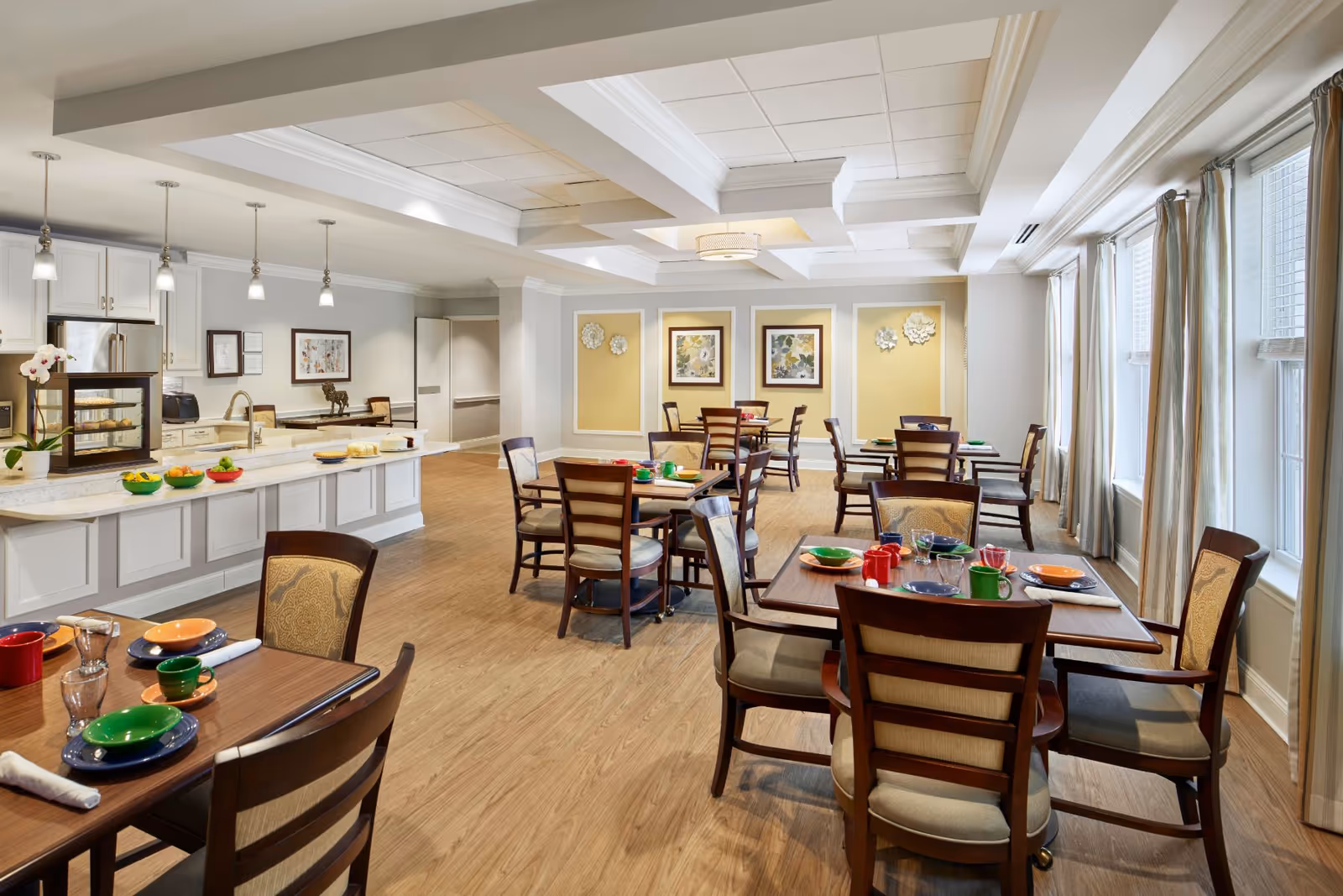 Bright dining room with multiple wooden tables and chairs, place settings, and a serving counter/kitchen area.