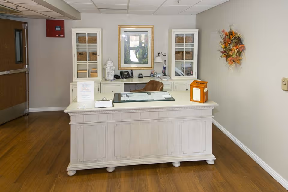 Reception desk area in a senior living facility with a white wooden desk, a brown chair, two white cabinets with glass doors containing baskets, a framed picture on the wall, a wall-mounted lamp, a decorative fall wreath, and a wooden floor.