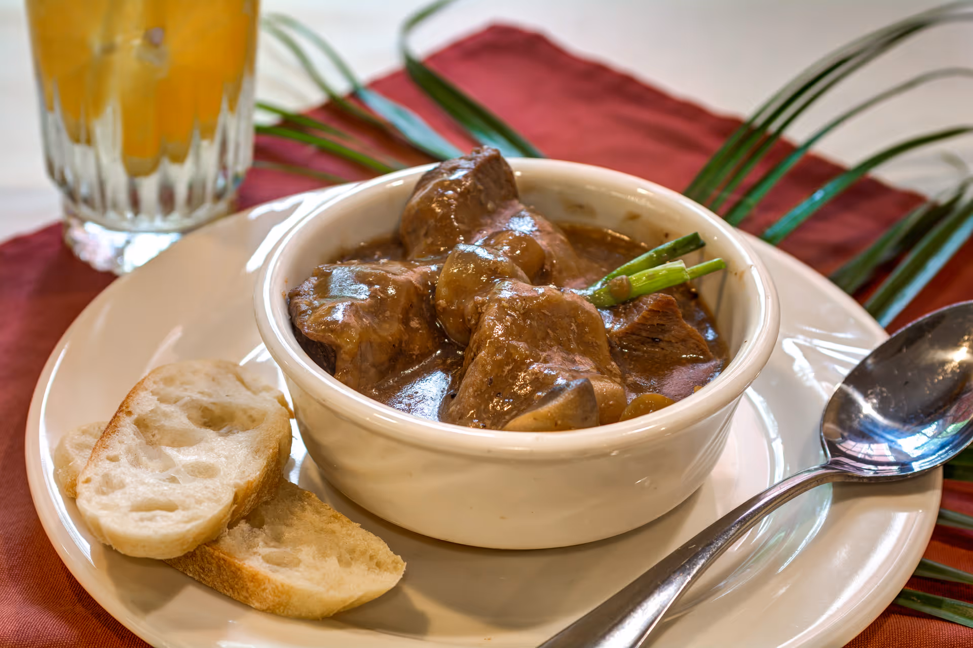 A bowl of beef stew with chunks of meat and vegetables garnished with green onions, served on a white plate with two slices of bread and a spoon. A glass of iced beverage is visible in the background on a red placemat.