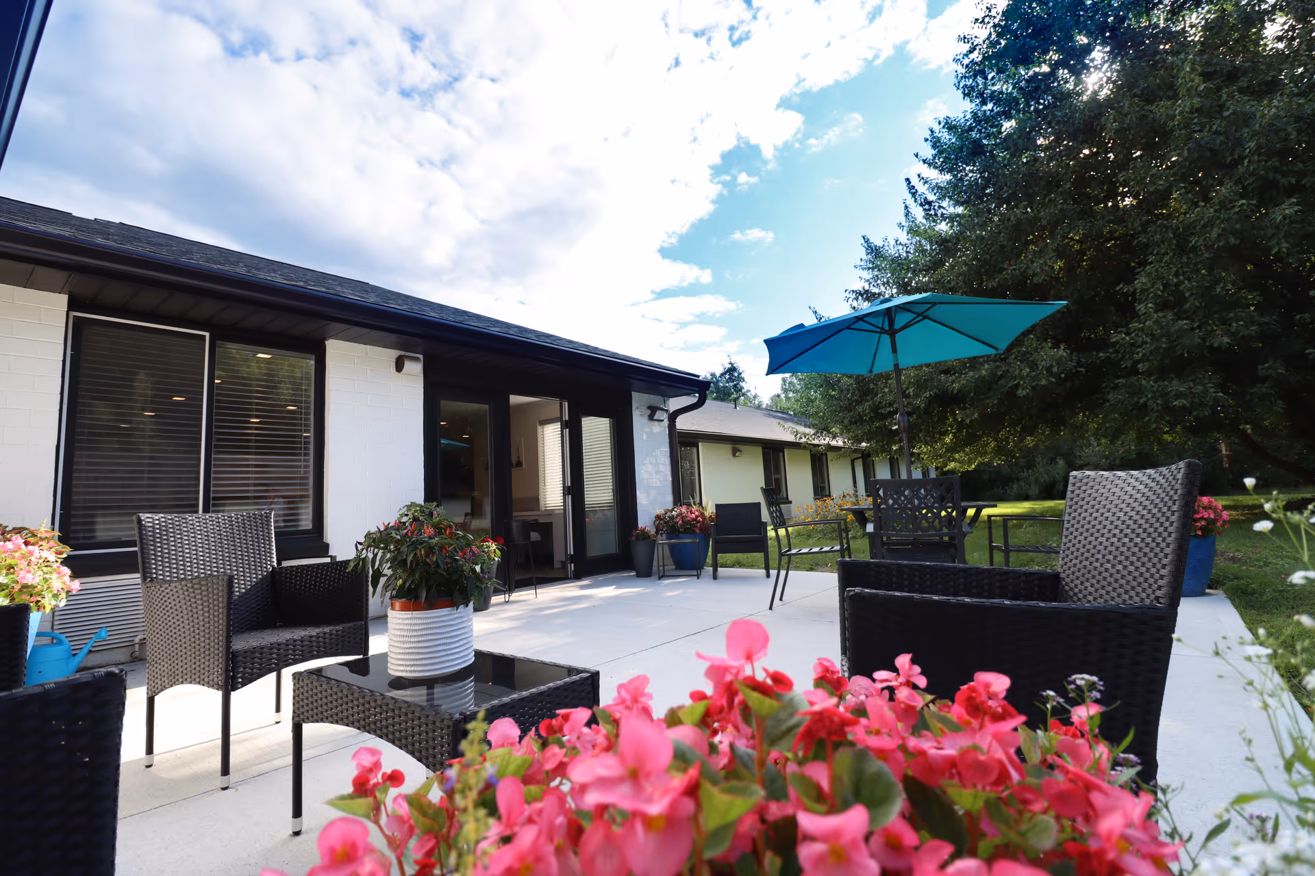 Outdoor patio area at Serenity Care Mid Valley with black wicker chairs and tables, a blue umbrella, potted plants, and pink flowers in the foreground. The patio is adjacent to a white building with large windows and a door, under a partly cloudy sky.