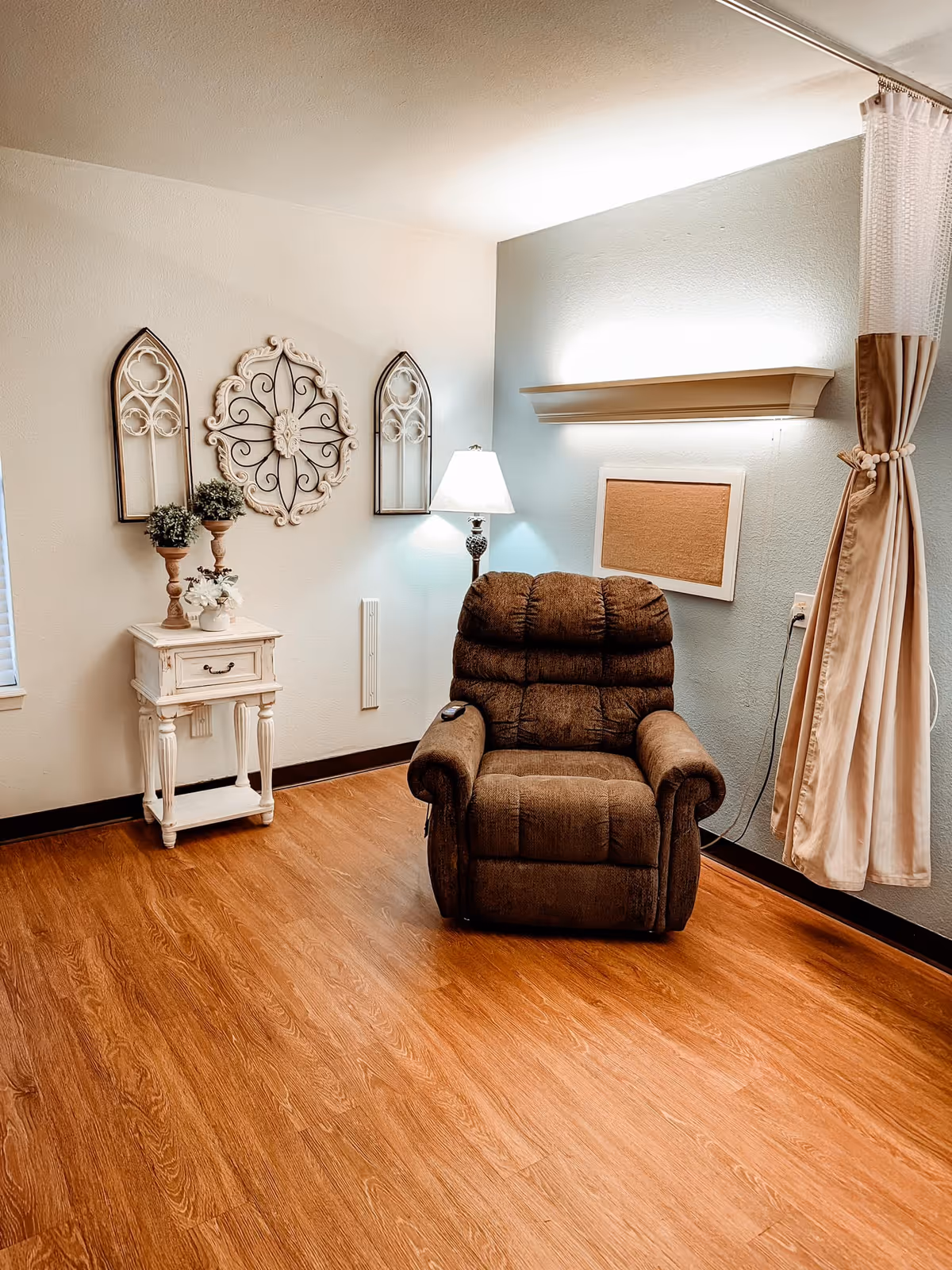 A cozy corner in a room featuring a brown recliner chair on a wooden floor. To the left of the chair is a small white side table with decorative plants and flowers. Above the table are three wall decorations with intricate designs. A floor lamp with a white shade is positioned behind the chair, and a beige curtain is tied back on the right side of the image. The walls are painted in light colors, with one wall having a light blue accent.