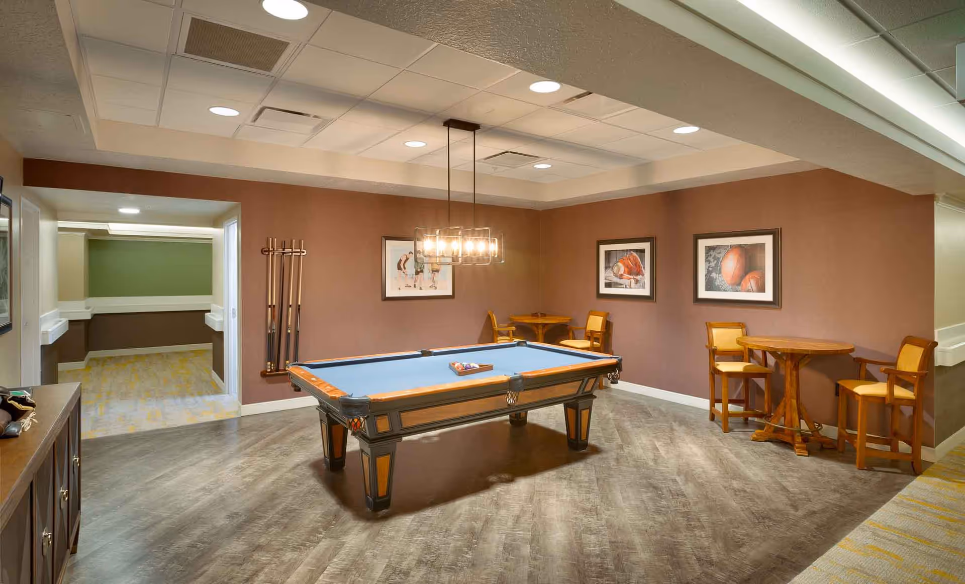 A recreational room with a pool table in the center, a hanging light fixture above it, and a set of pool cues mounted on the wall. There are two small round tables with chairs against the walls, and framed pictures hanging on the brown walls. The floor is wood-patterned, and there is an open doorway leading to a hallway with green and brown walls and carpeted floor.